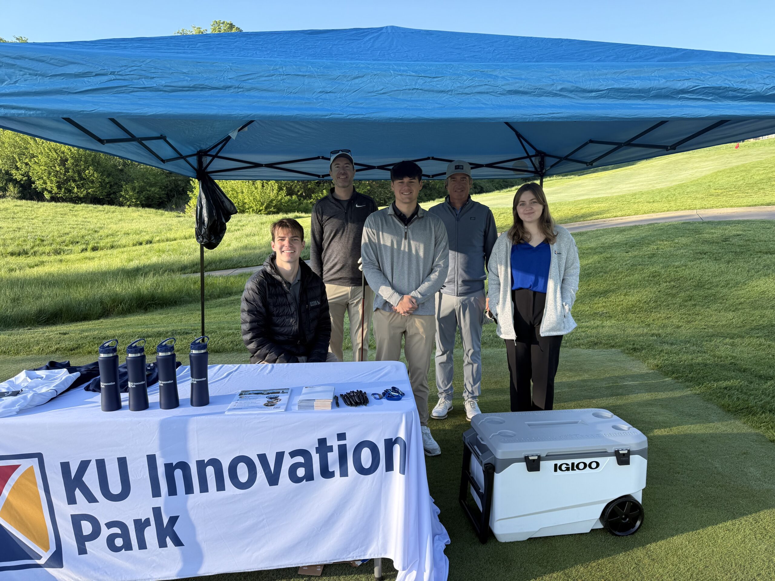 Sand Trap pic Five people pose together under a blue canopy tent on a golf course, standing behind a KU Innovation Park table. The table is covered with branded materials including navy water bottles, pens, and brochures. A large Igloo cooler sits nearby on the grass. The group is casually dressed for an outdoor event, with a sunny, green landscape in the background.